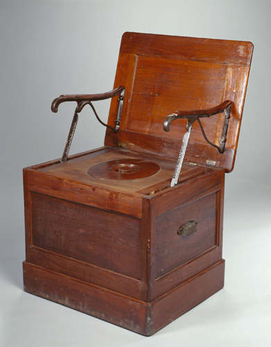 A wooden invalid chair shaped like a box with handles has a built-in chamber pot.